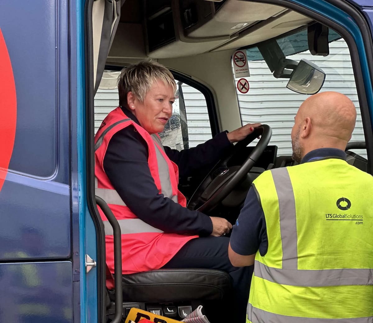 A woman behind the wheel of a truck, with a man stood outside talking to her